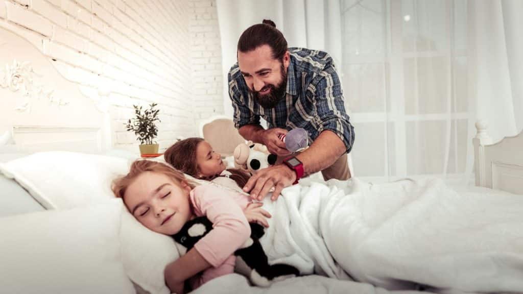 A smiling bearded man tucks two young girls into a bed.