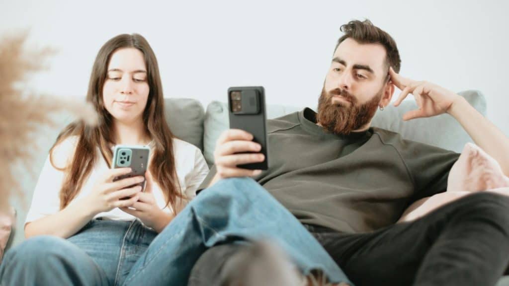 A couple sits on a couch, both silently looking down at their cell phones.