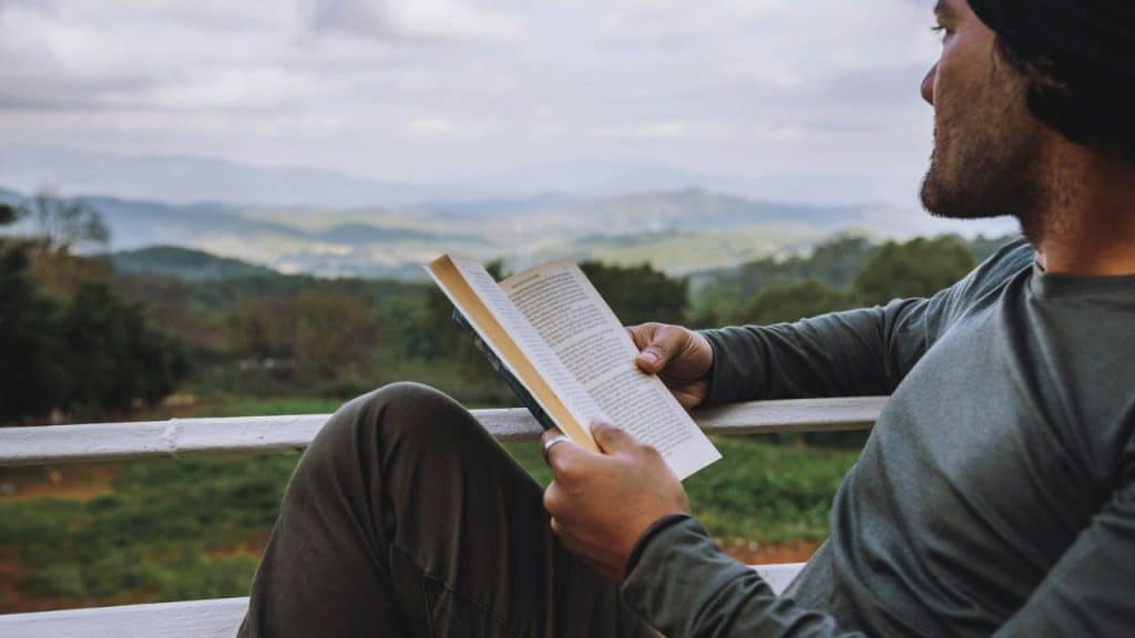 A man sits outdoors reading a book, overlooking a green valley and mountains.