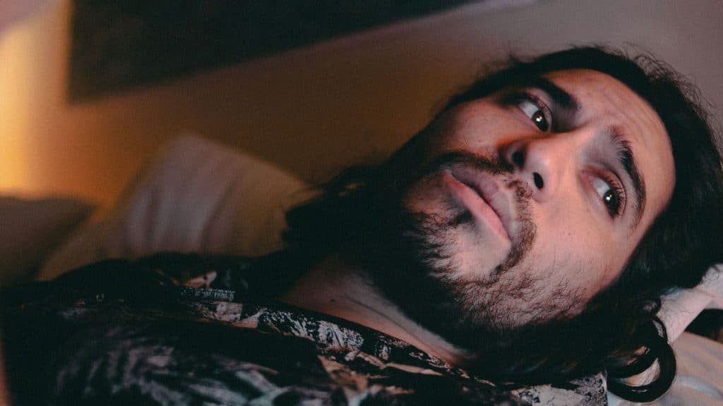 A bearded man with long hair lies in bed, looking away with an intense expression.