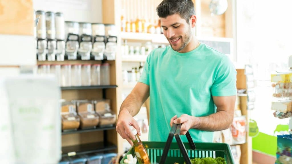 A smiling man in a teal shirt is placing an item into a green shopping basket.