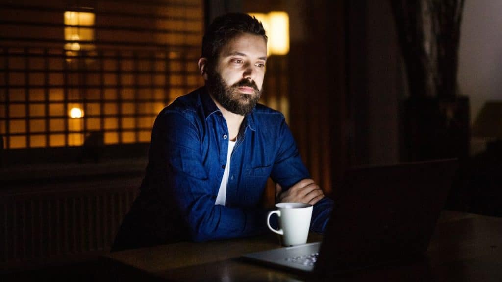 A bearded man is working late on a laptop in a dimly lit room.
