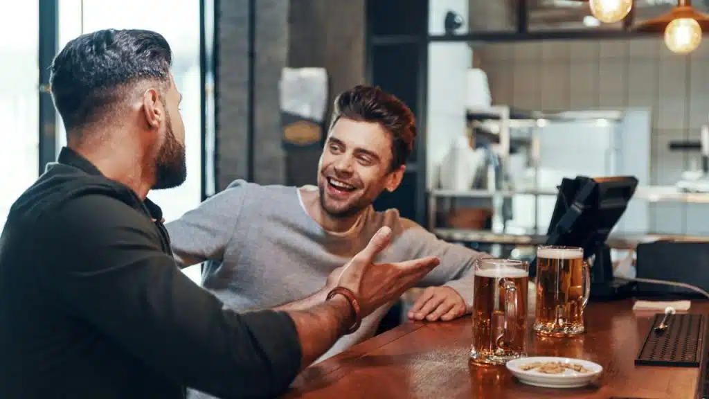 Two men sitting at a bar, one laughing and the other gesturing with his hand, with two mugs of beer on the counter.