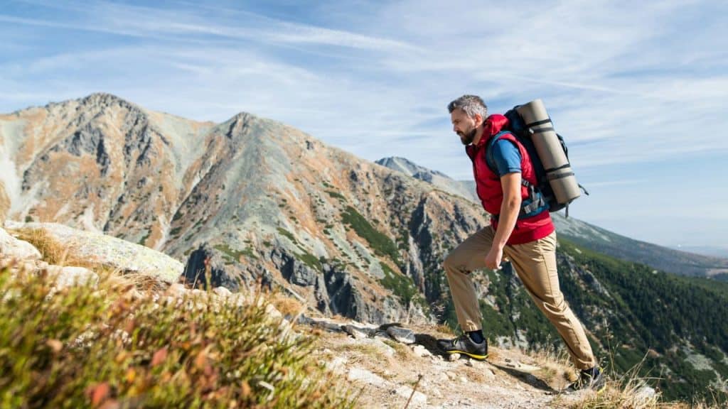 Middle-aged man hiking uphill on a mountain trail with a backpack and sleeping mat.
