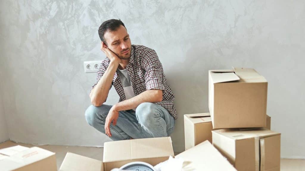 Man sits glumly on his haunches in a room with cardboard boxes.