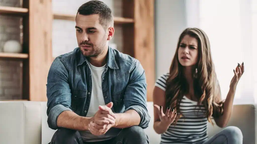 A man sits looking away somberly while a woman gestures angrily at him on a couch.