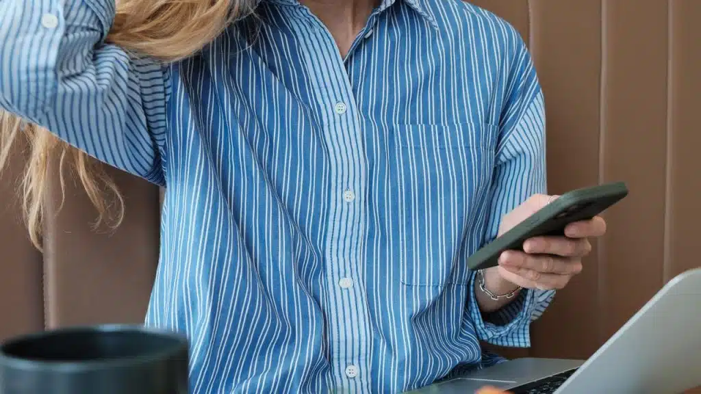 A person in a blue striped shirt holding a phone while sitting at a table with a laptop.