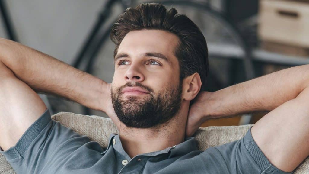 Bearded man relaxing on a couch, looking up and to the right, hands behind his head.