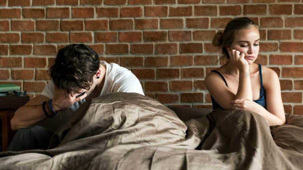 Man and woman sitting apart in bed, covering their faces or resting their heads on their hands.