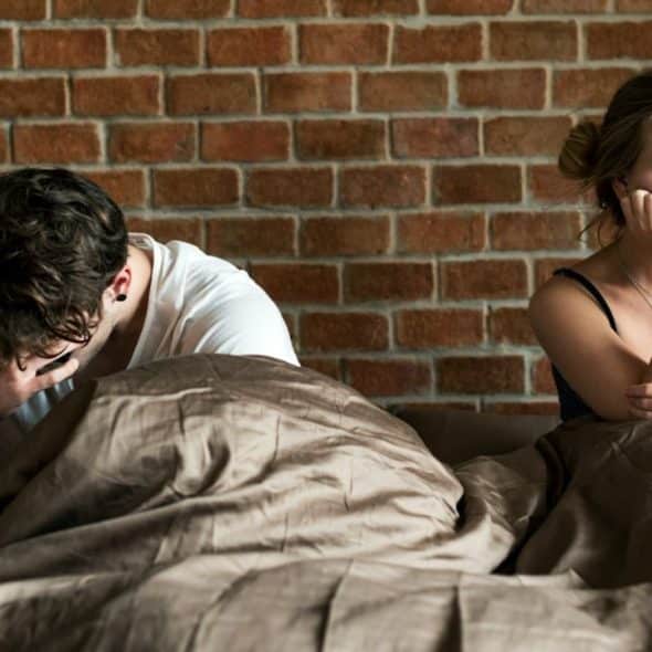 Man and woman sitting apart in bed, covering their faces or resting their heads on their hands.