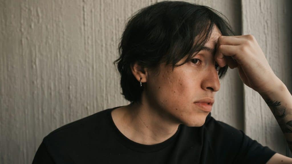 Young man with dark hair in a black shirt, resting his hand on his forehead, looking away with a pensive expression.