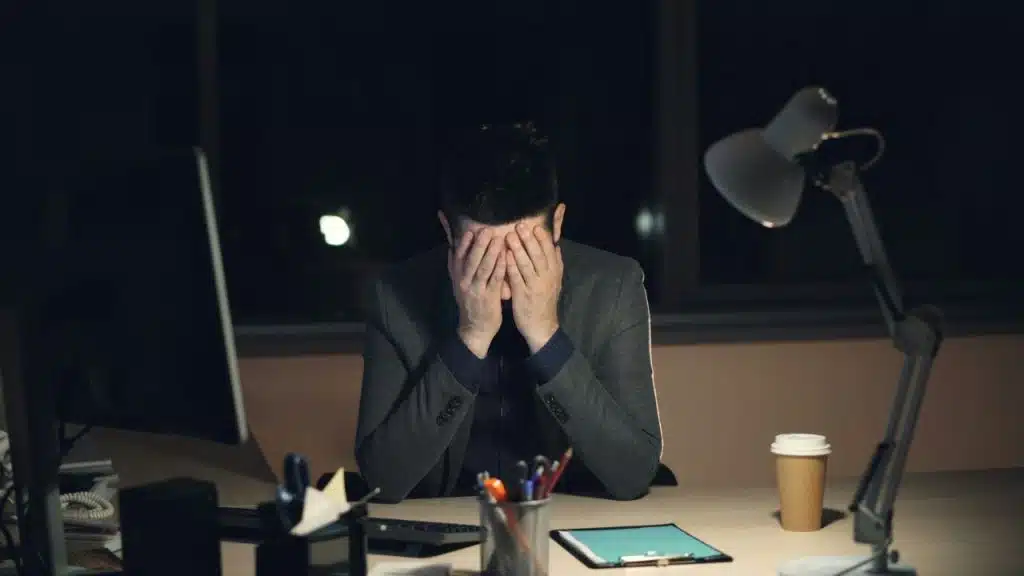 A man sits at a desk in the dark, covering his face with his hands in frustration.