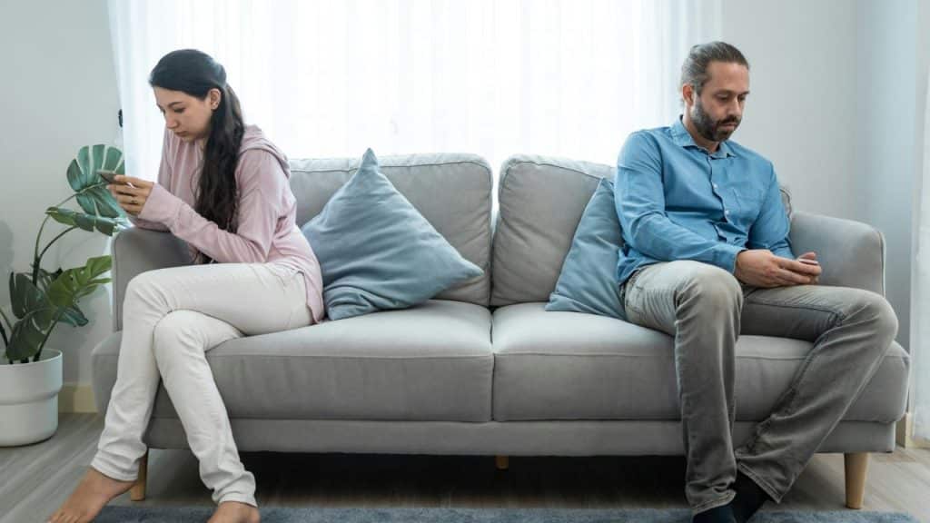 A man and woman sit far apart on a couch with a pillow between them.