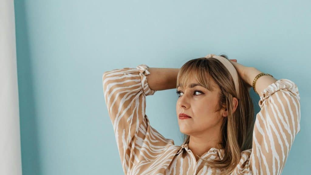 A woman leaning back with her hands on her head against a blue wall.