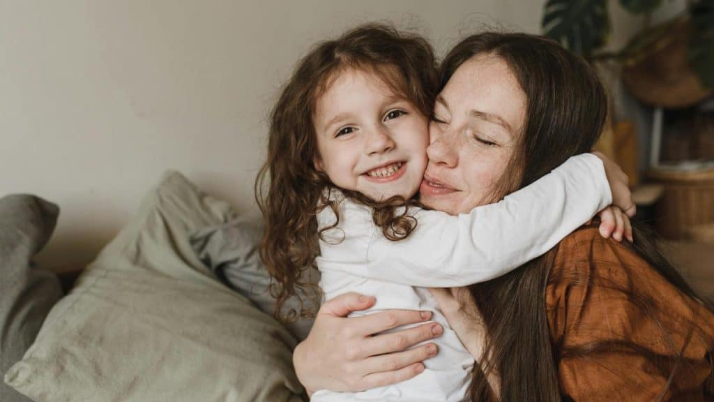 A mother and daughter hugging and smiling on a couch.