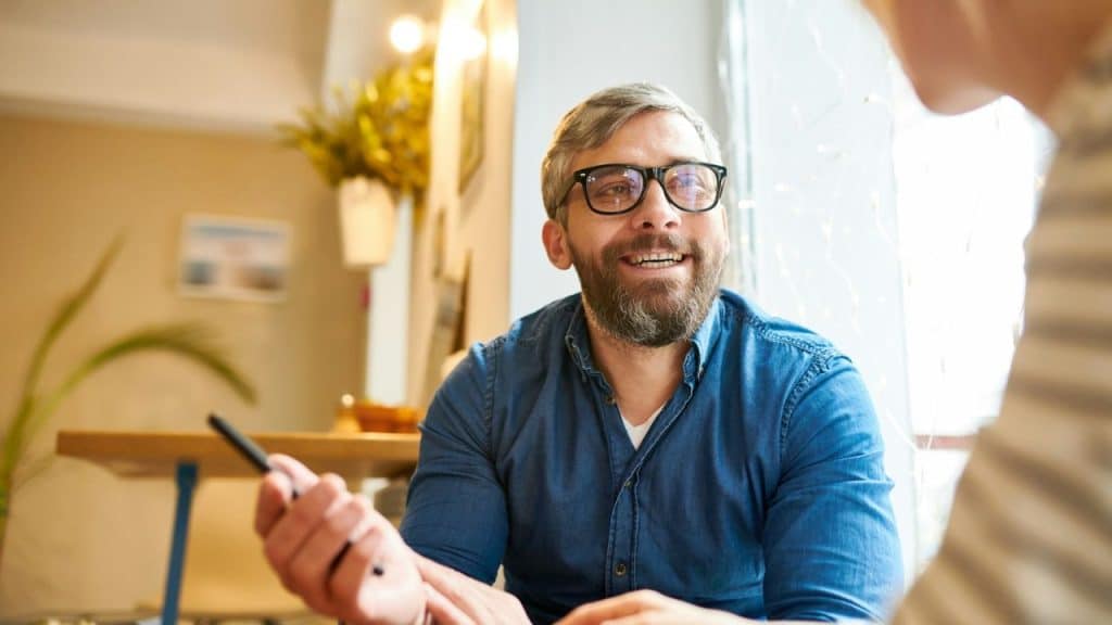 Bearded man in a denim shirt and glasses smiles while holding a pen indoors.