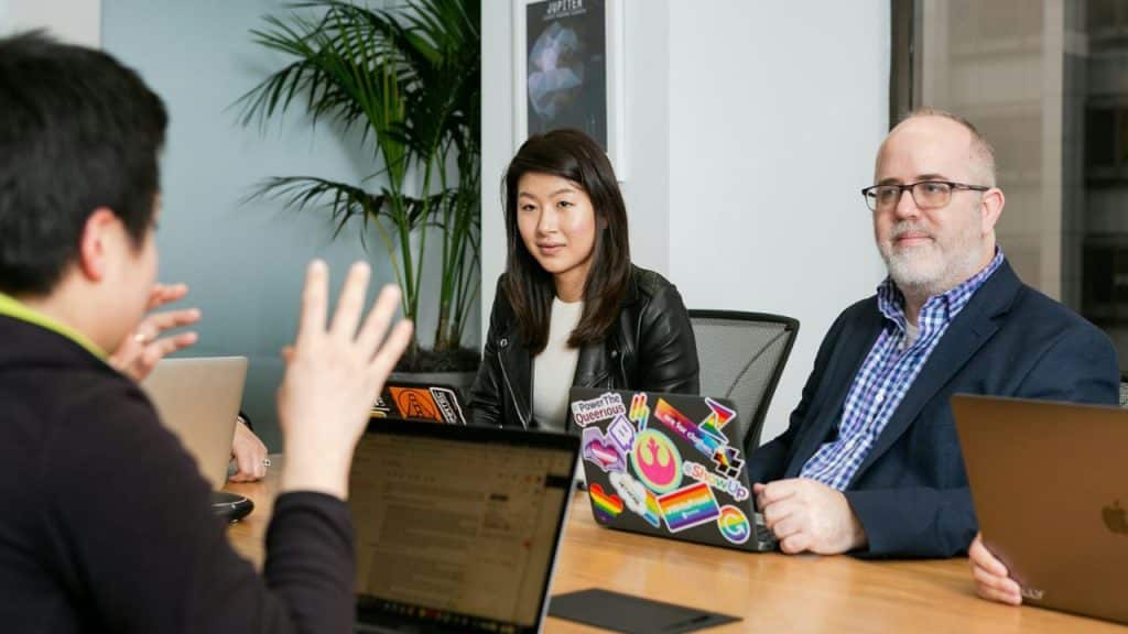 Three people are having a discussion around a table with laptops in an office.