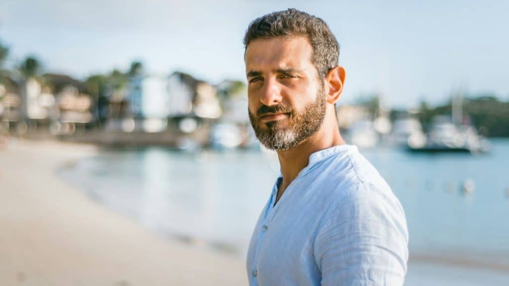 Man with a beard in a light blue shirt looking at the camera on a sunny beach.