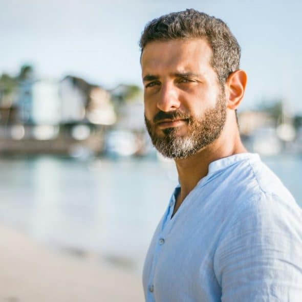 Man with a beard in a light blue shirt looking at the camera on a sunny beach.