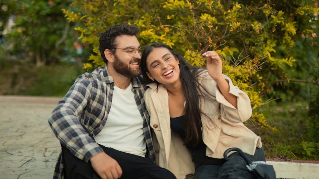 A happy couple sitting in a park with the woman leaning on the man’s shoulder.