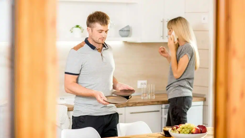 A man is holding plates in a kitchen while a woman is talking on a cell phone in the background.