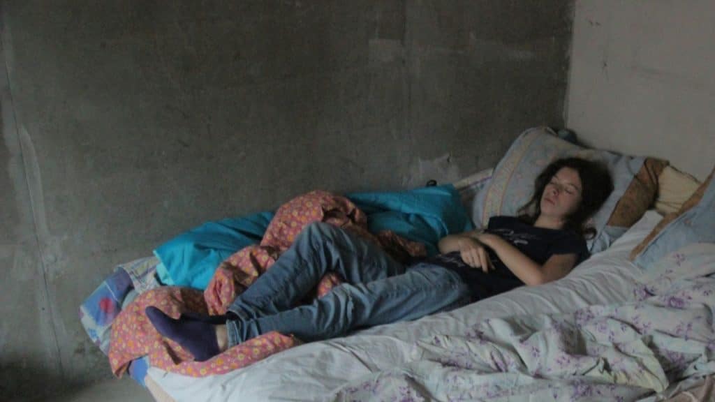 A woman sleeping on a mattress pad in a dark, empty room with grey, rough walls.