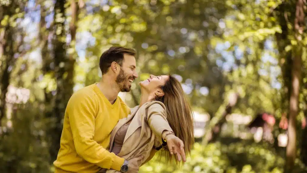 A joyful couple embracing and laughing in a park, with the man holding the woman as she leans back playfully, surrounded by lush green trees.