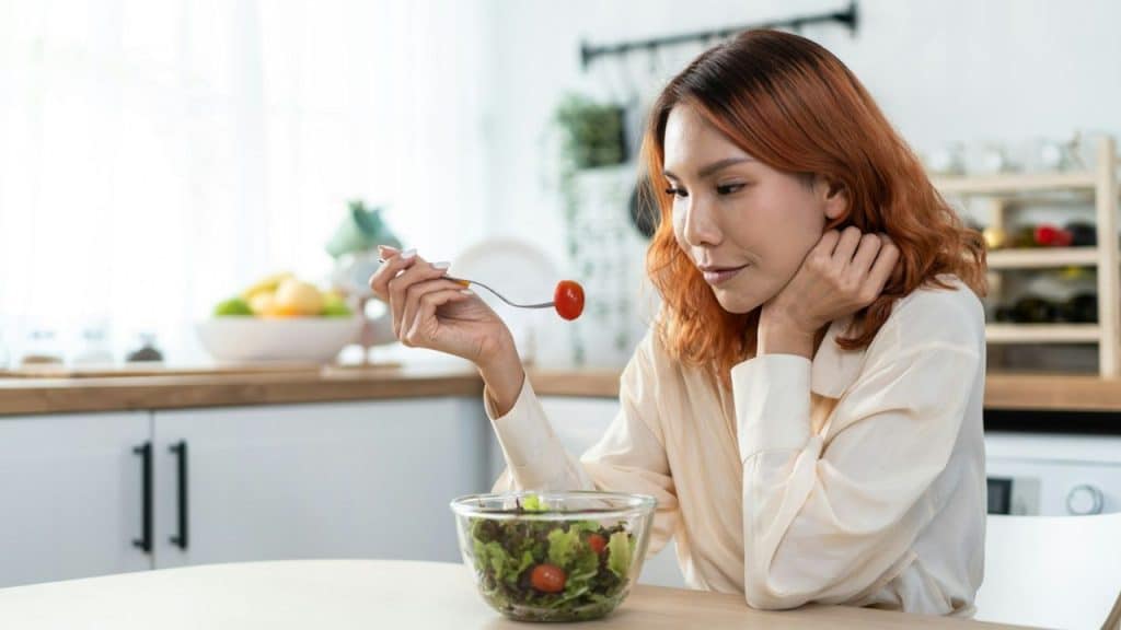 A woman with auburn hair sits at a light wood table in a kitchen, looking at a cherry tomato on a fork over a salad bowl.