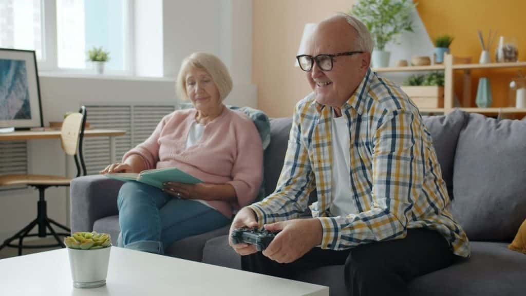 An older man is smiling while playing a video game on a couch, and an older woman is reading a book next to him.