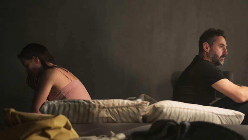 A man and woman are sitting on a bed facing away from each other in a dark room after an argument.