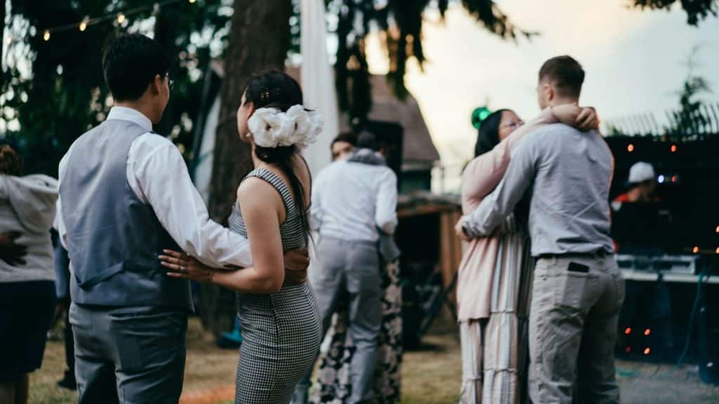 Two couples dance closely at an outdoor evening gathering, with the woman on the left wearing white flowers in her hair.