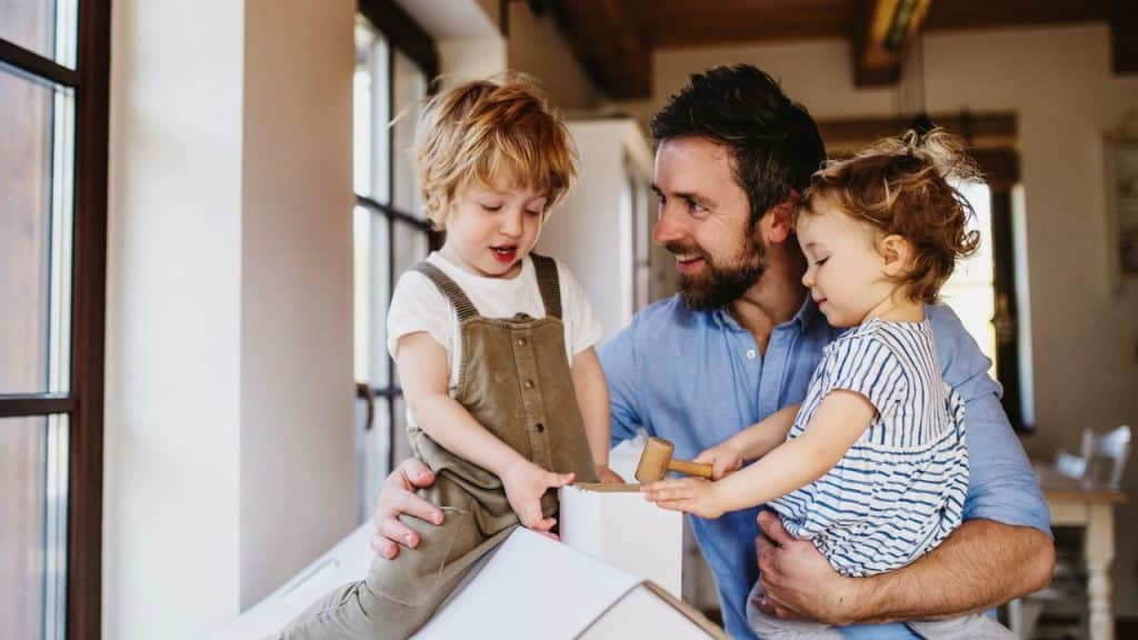 Father smiles at his two young children playing with a small wooden hammer.
