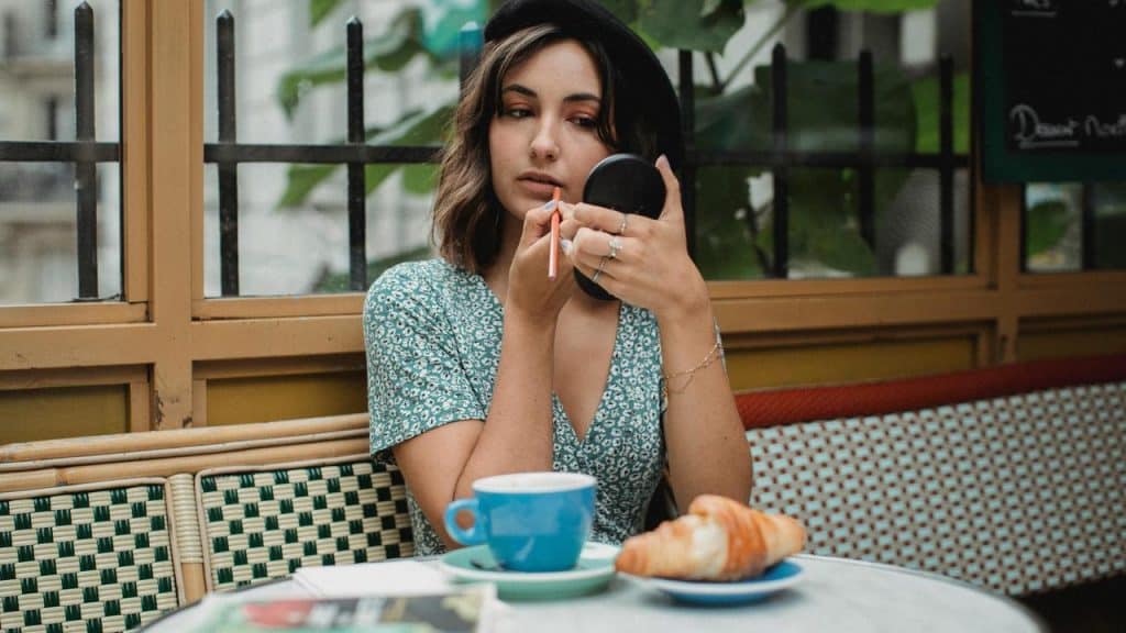 A woman applying lipstick at a café table with coffee and a croissant.