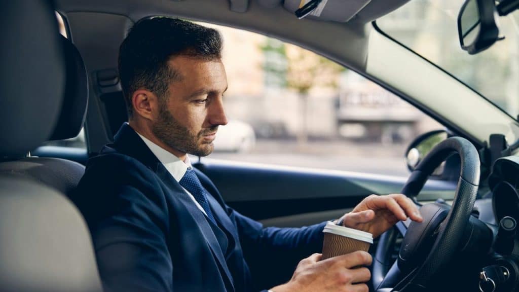 Man in a dark suit and tie sitting in a car, holding a coffee cup.