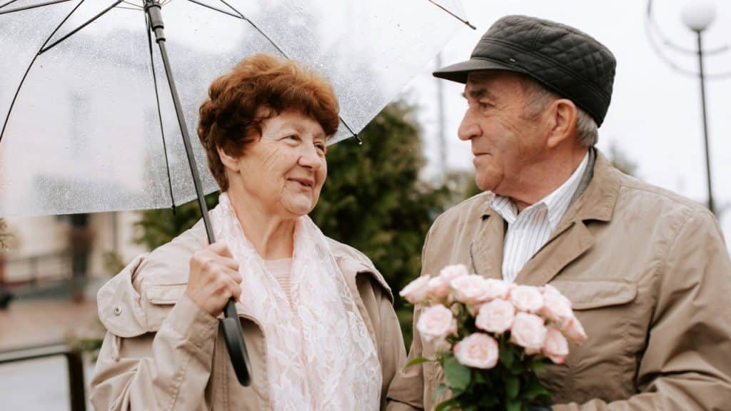 Older couple outdoors; man holds pink roses, woman holds a clear umbrella.