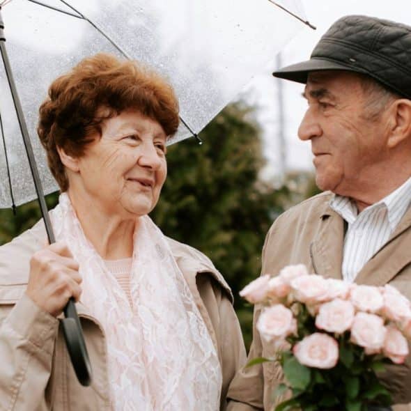 Older couple outdoors; man holds pink roses, woman holds a clear umbrella.