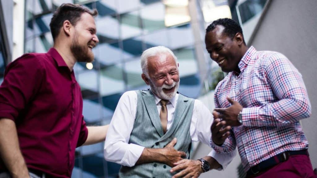 Three men of different ages and races are standing outside a modern building and laughing.
