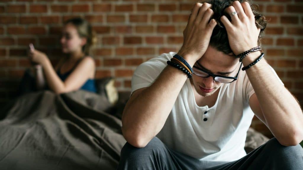 A distressed man with glasses and bracelets sits on a bed while a woman in the background looks at her phone.