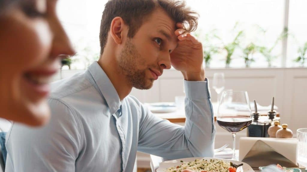 Man sits at a restaurant table, head resting on his hand, looking away from a smiling person.