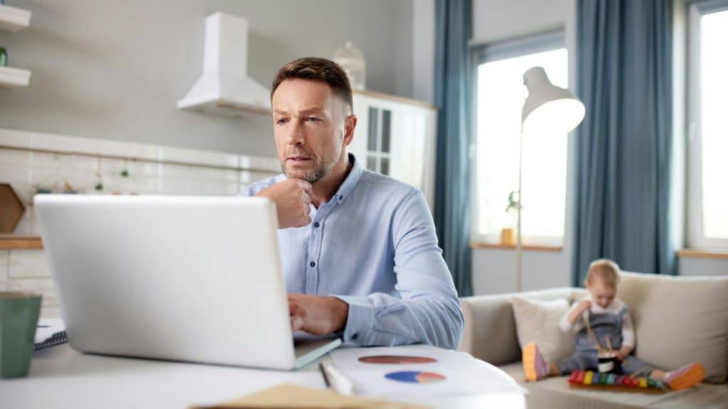 Man sits at a laptop with papers, looking focused, while a baby plays on a couch behind him.