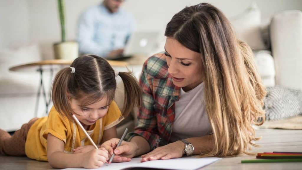 A woman in a plaid shirt is helping a young girl draw with a pencil on the floor, while a man is visible working in the blurred background.
