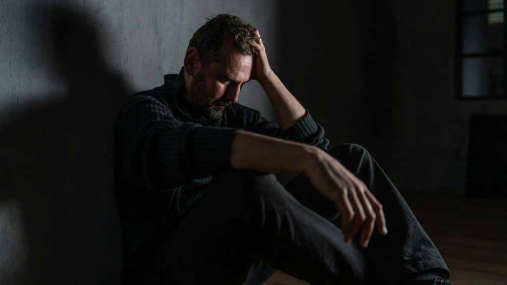 A man is sitting on the floor in a dimly lit room, with his head down and hand touching his forehead.