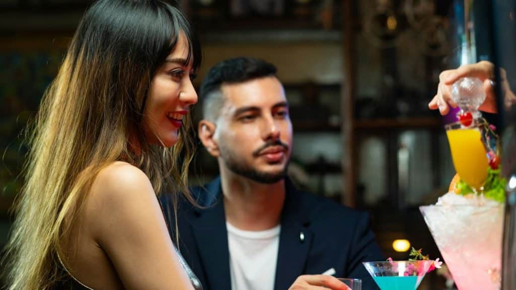 Woman smiles next to a man at a bar while a bartender prepares a colorful drink.