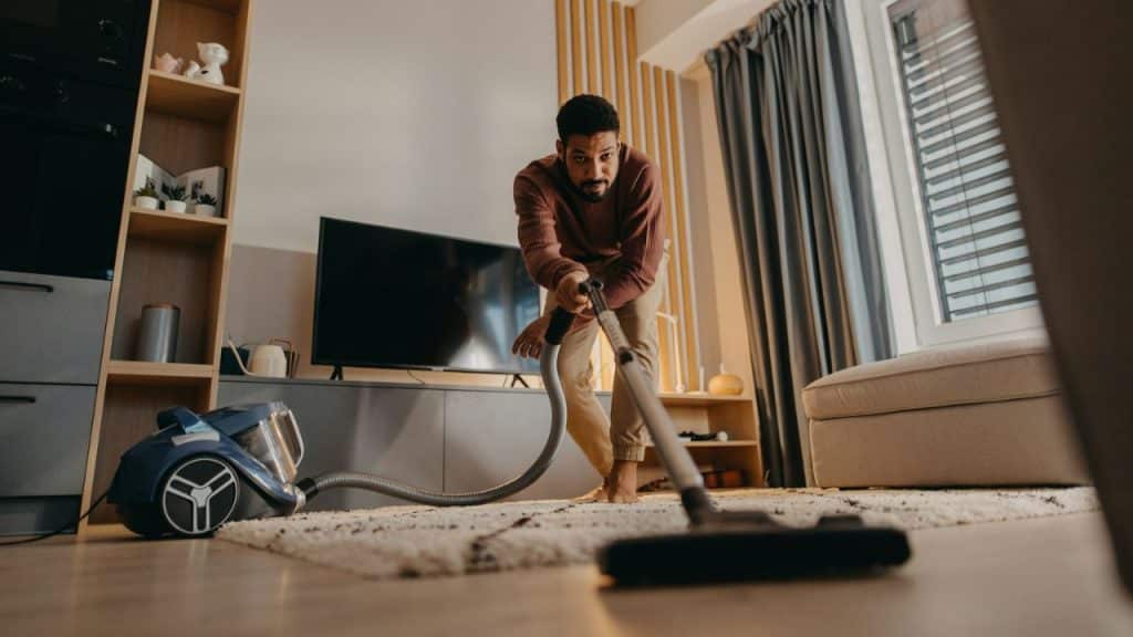 Man vacuuming a shaggy rug in a living room, looking directly at the camera.