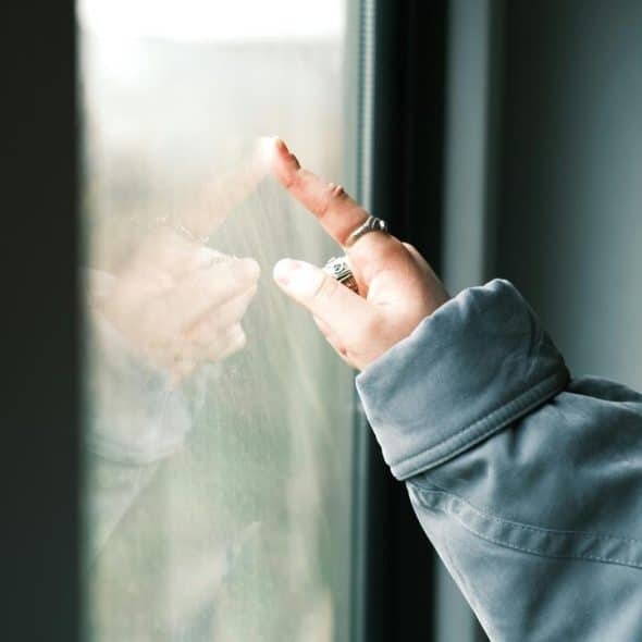 A person’s hand wearing rings touches a window, showing its reflection on the glass.