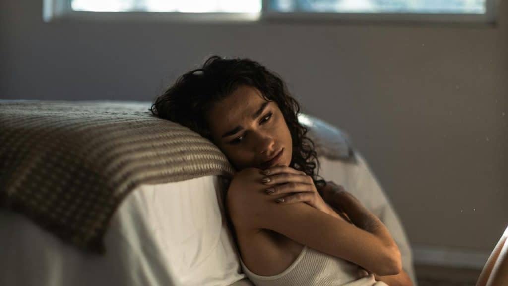 A woman sitting on the floor leaning against a bed, looking sad.