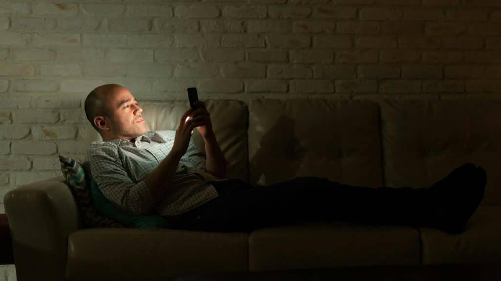 A man lying on a couch in a dark room looking at his phone.