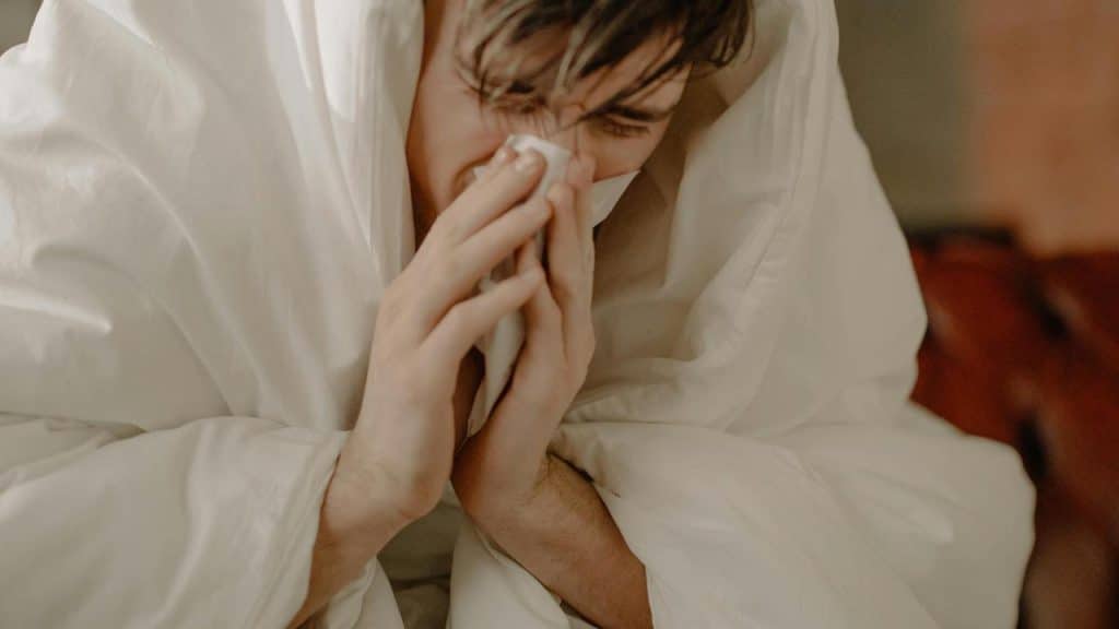 A person wrapped in a blanket blowing their nose with a tissue.