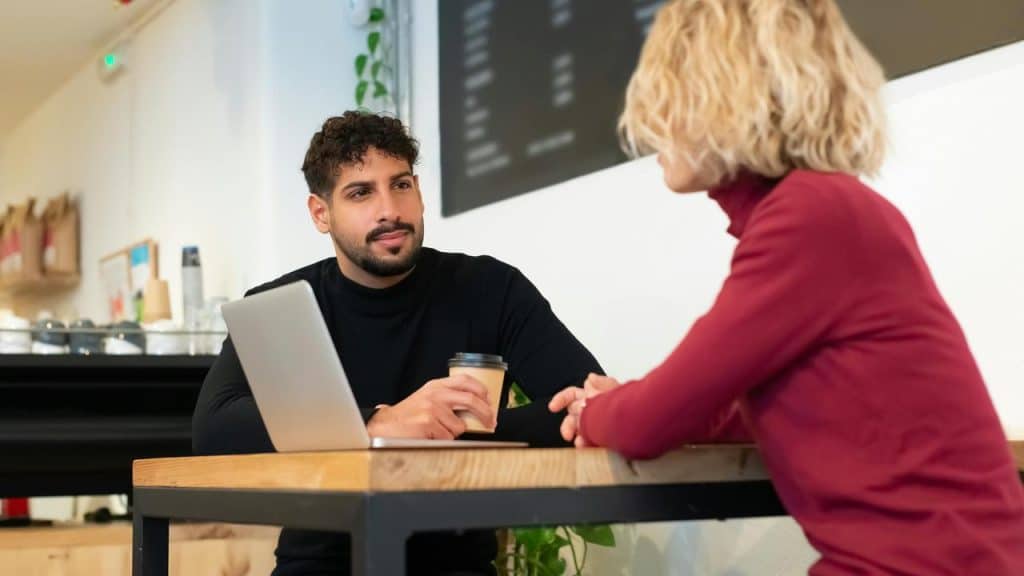 A man and woman sitting at a café table talking with a laptop and coffee cup.