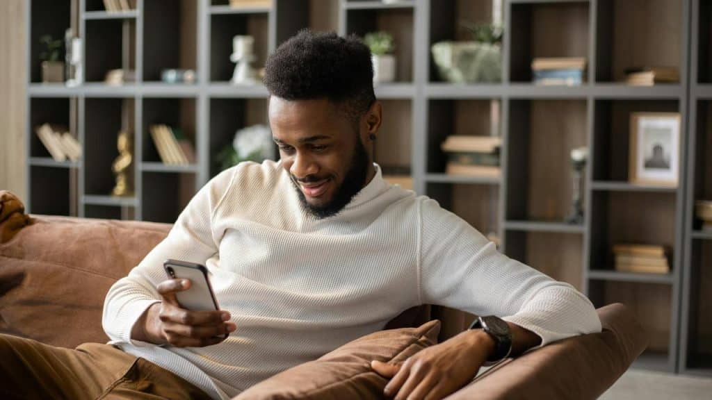 A man sitting on a couch smiling while looking at his smartphone.